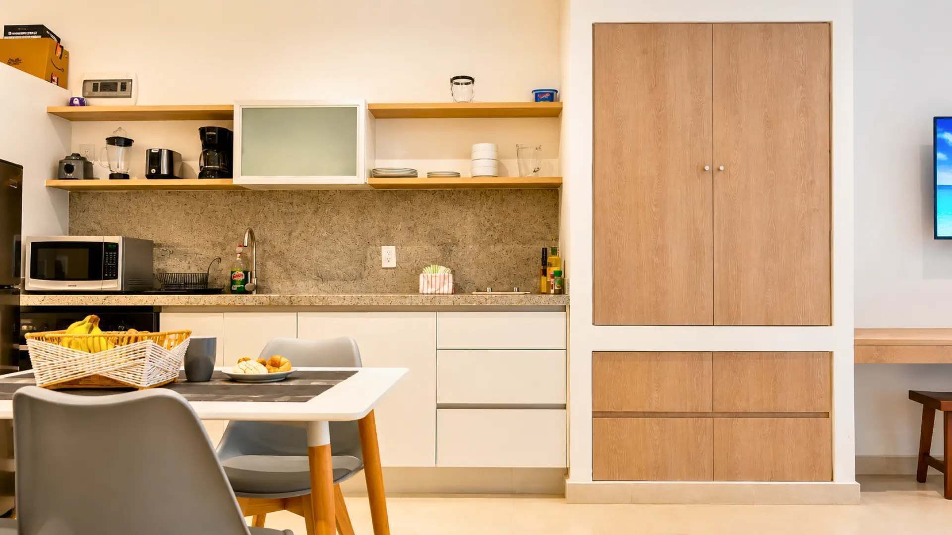 Une cuisine compacte équipée d’un micro-ondes, d’un grille-pain et d’une machine à café sur le plan de travail, d’une table à manger avec des chaises et d’une grande armoire en bois.