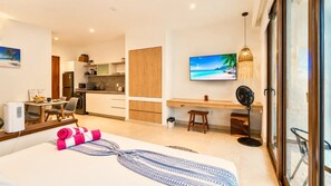 Ein modernes Hotelzimmer mit Bett, Schreibtisch, Küchenzeile und einem Fernseher, der eine Strandansicht zeigt.