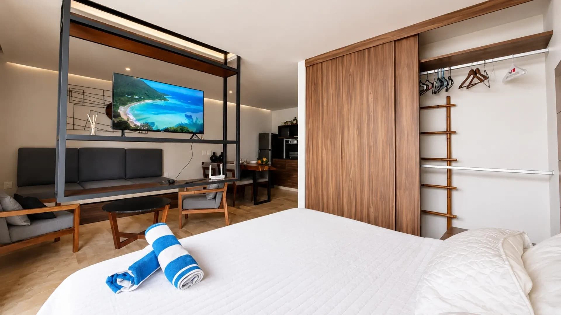 A modern hotel room with a large bed, a TV mounted on a wall, a seating area with a sofa and chairs, and a wooden wardrobe.