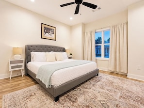 A bedroom with a large bed, a ceiling fan, a window with curtains, and a framed picture on the wall.