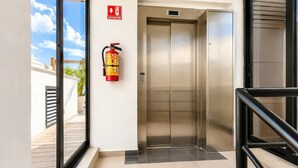 Een lift met een brandblusser aan de muur, een heldere glazen deur en een metalen handgreep.