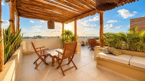 Una pergola in legno con un cesto intrecciato, un tavolo di legno con due sedie, un divano con cuscini e piante in vaso su una terrazza sul tetto con vista sulla città.