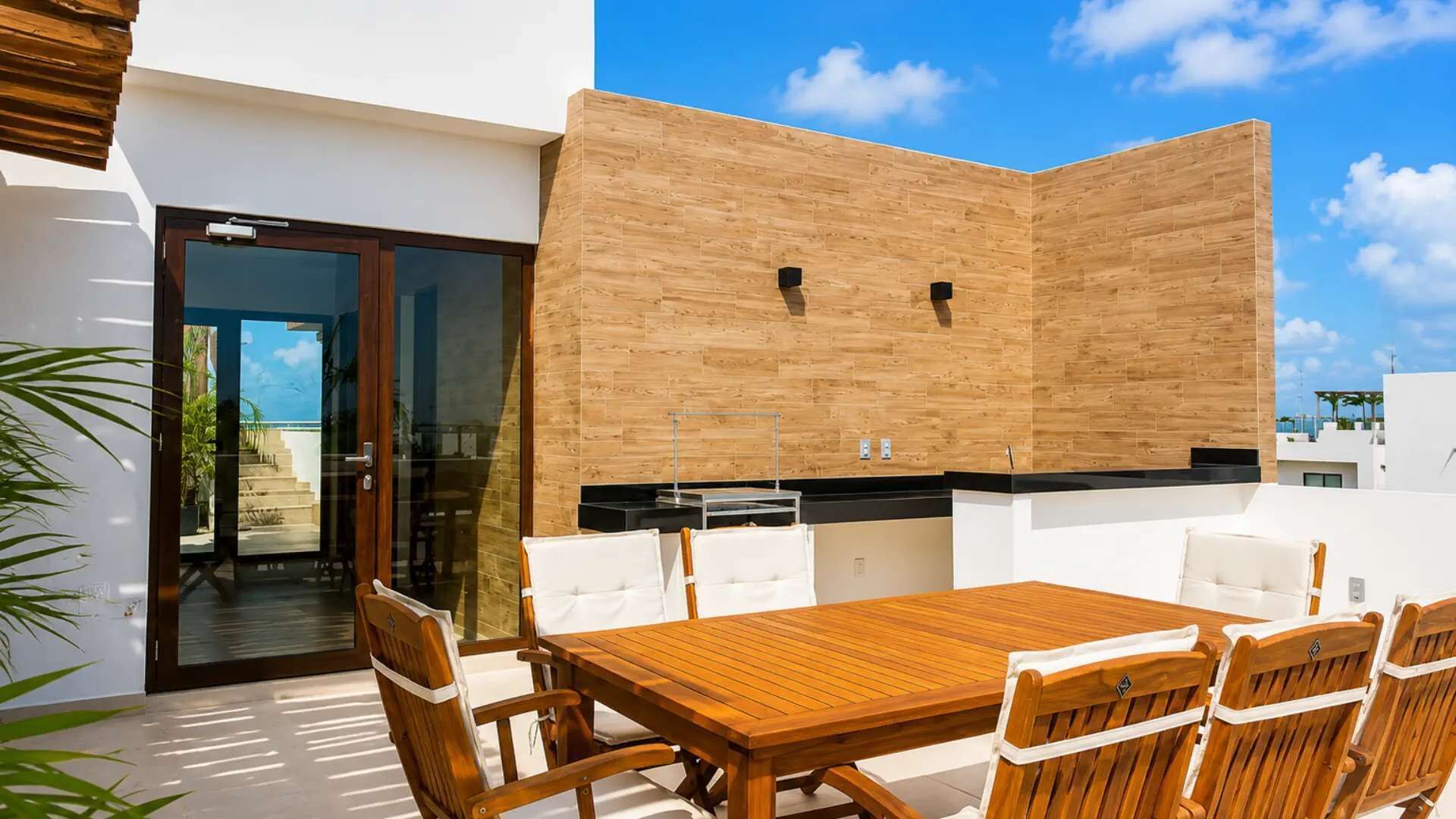 Een terras met een houten tafel en stoelen, een glazen deur die uitkomt bij een zwembad, en een houten wand met twee speakers.