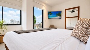 A bedroom with a large bed, a view of the city through a window, a ceiling fan, and a painting of a beach. - Dreamy Rooftop Pool Studio near Beach · Cosmo 42 (Playa del Carmen)