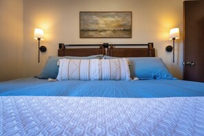 Una cama bien tendida con una colcha estampada, dos lámparas fijadas a la pared y un cuadro sobre la cabecera.