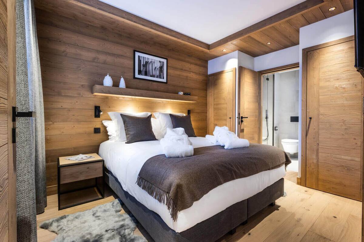 A modern bedroom with a wooden headboard, a large bed with white and brown bedding, a nightstand, and a wooden wardrobe.
