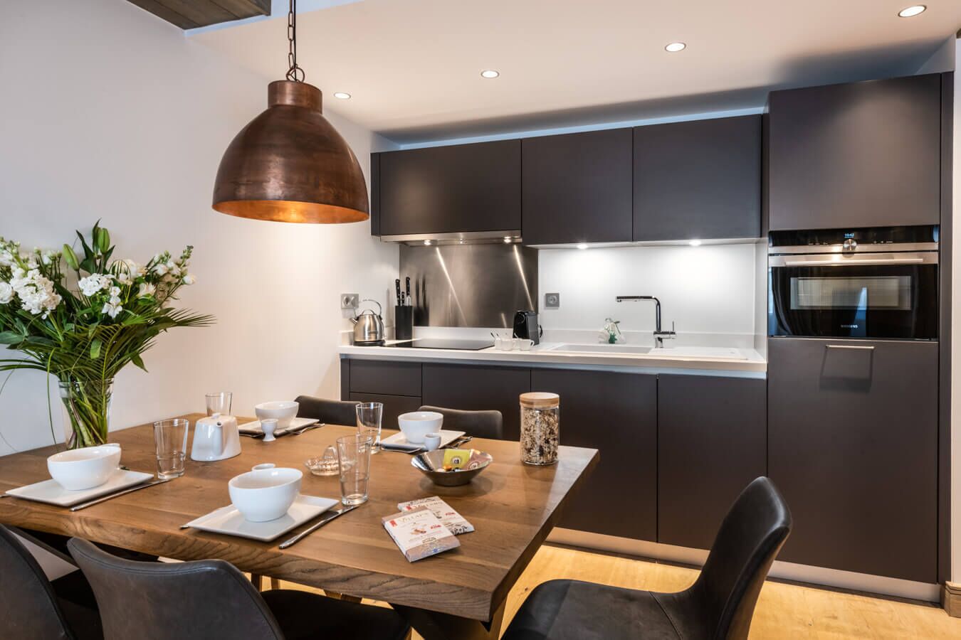 Cocina moderna con armarios oscuros, electrodomésticos de acero inoxidable y una mesa de comedor de madera para cuatro.