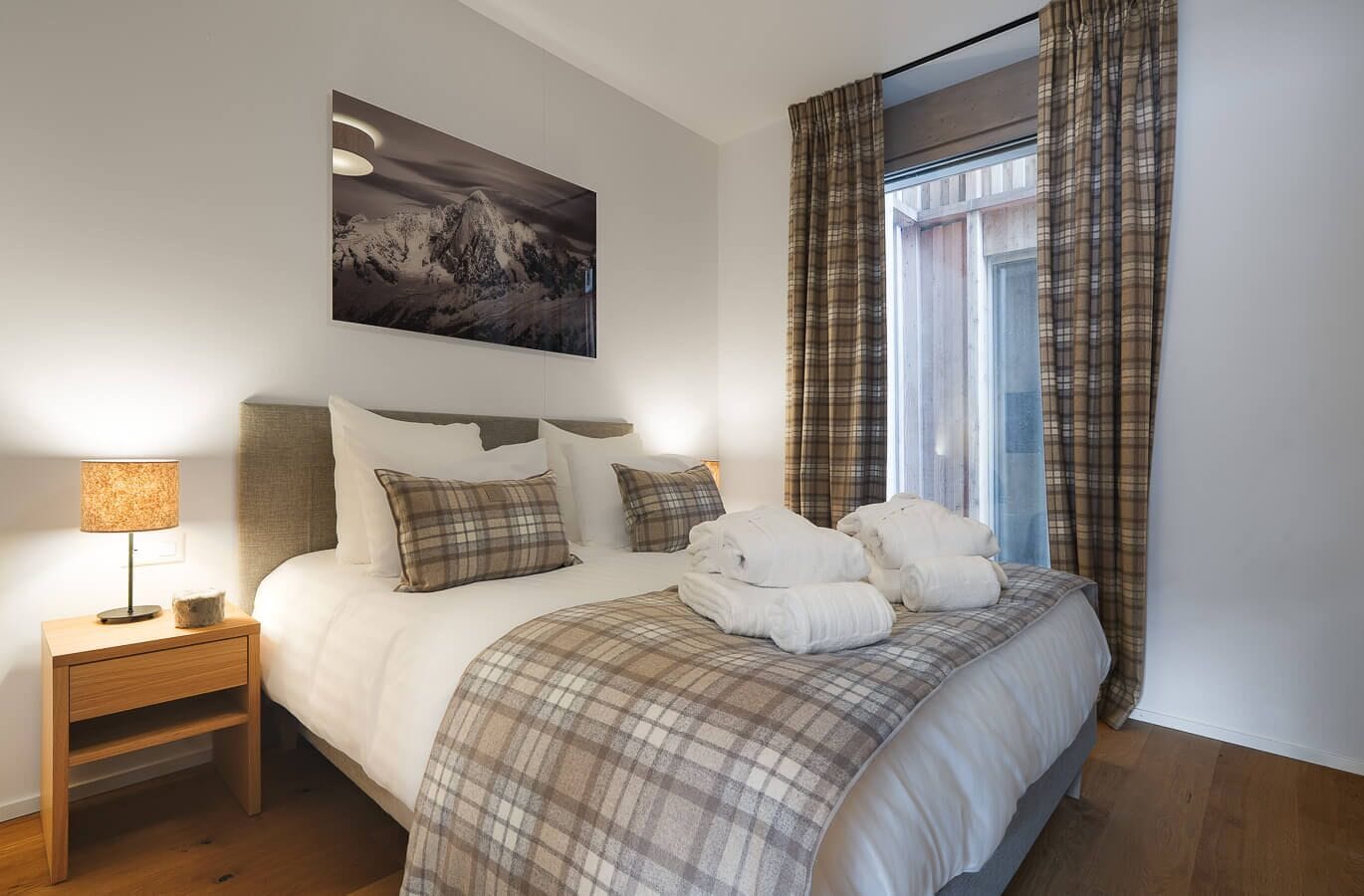 A hotel room with a bed, a nightstand, a lamp, a painting, and plaid curtains.