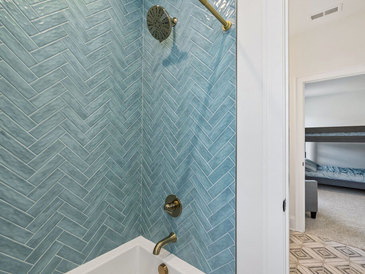 Une salle de bain avec un carrelage disposé en motif «chevrons», une douche dotée d’un robinet doré et une porte visible donnant sur une autre pièce.