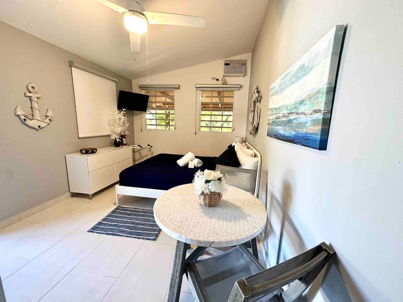 A bedroom with a bed, a bedside table, a chair, a small round table, a ceiling fan, a window, and a painting on the wall.