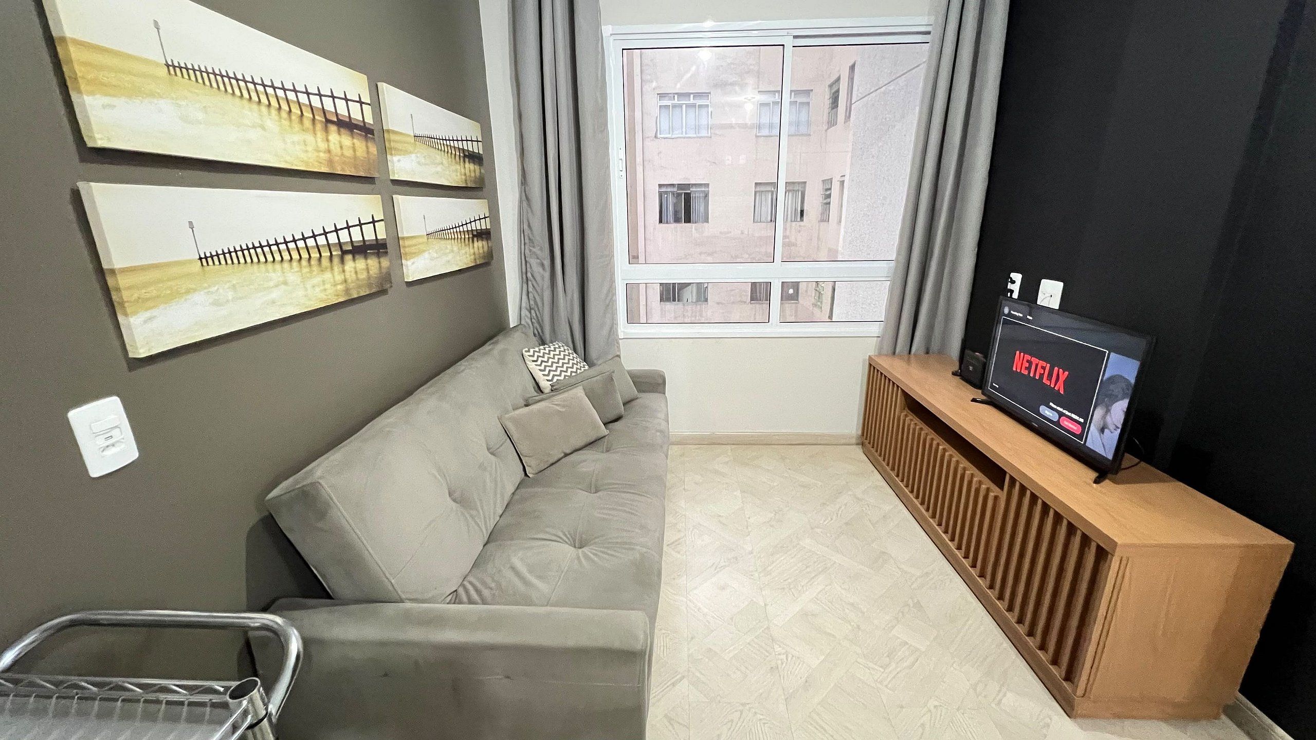A living room with a sofa, a wooden console table with a TV displaying 'NETFLIX', and a window with curtains.
