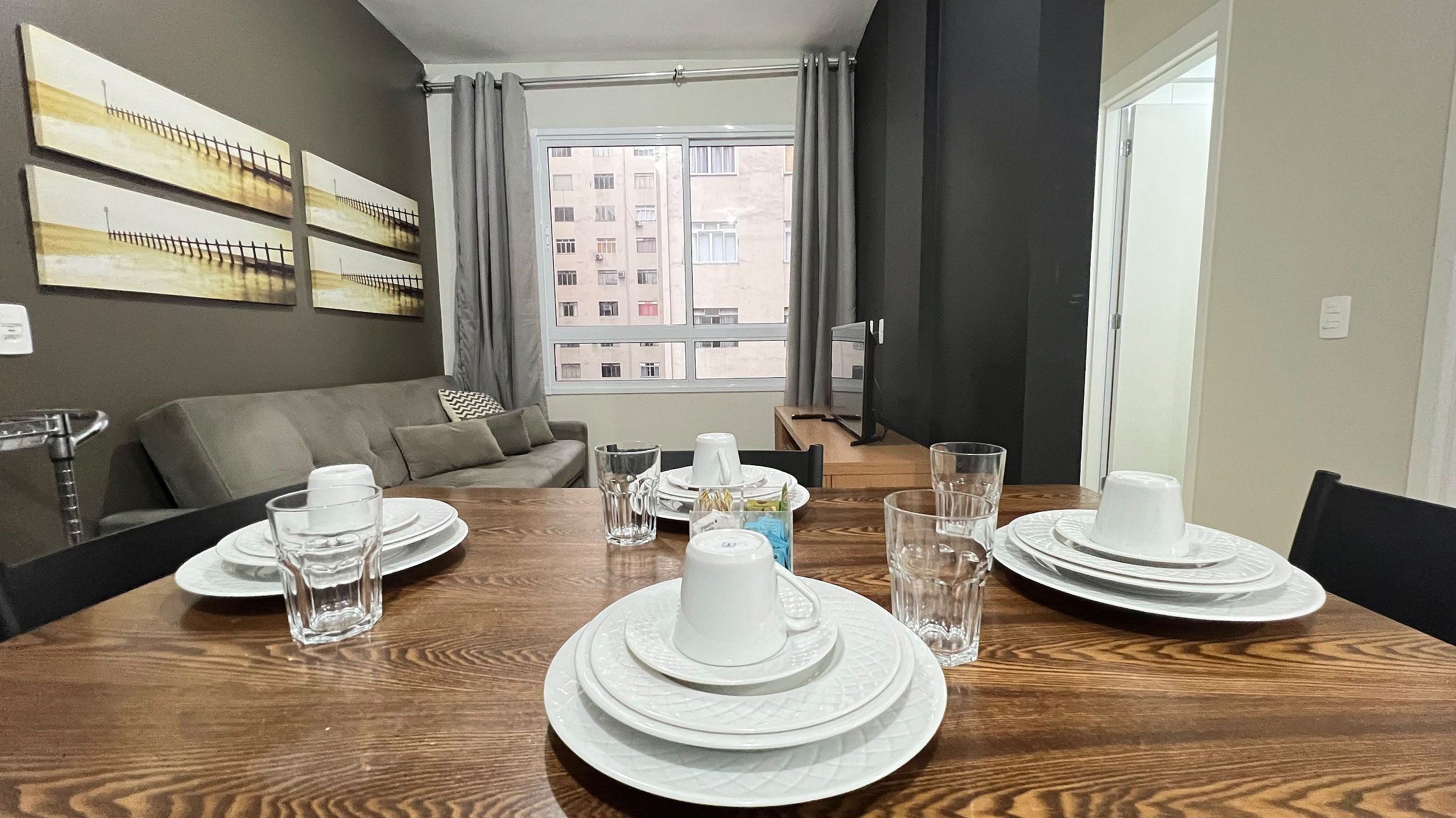 A dining area with a wooden table set for four, a sofa, and a view of buildings through a window.