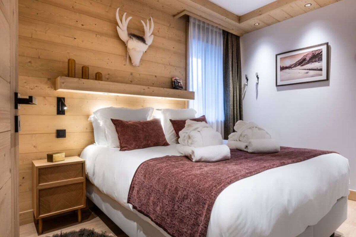 A bedroom with a wooden ceiling, a bed with white bedding and a red blanket, a mounted deer head, a nightstand, and a fur rug.