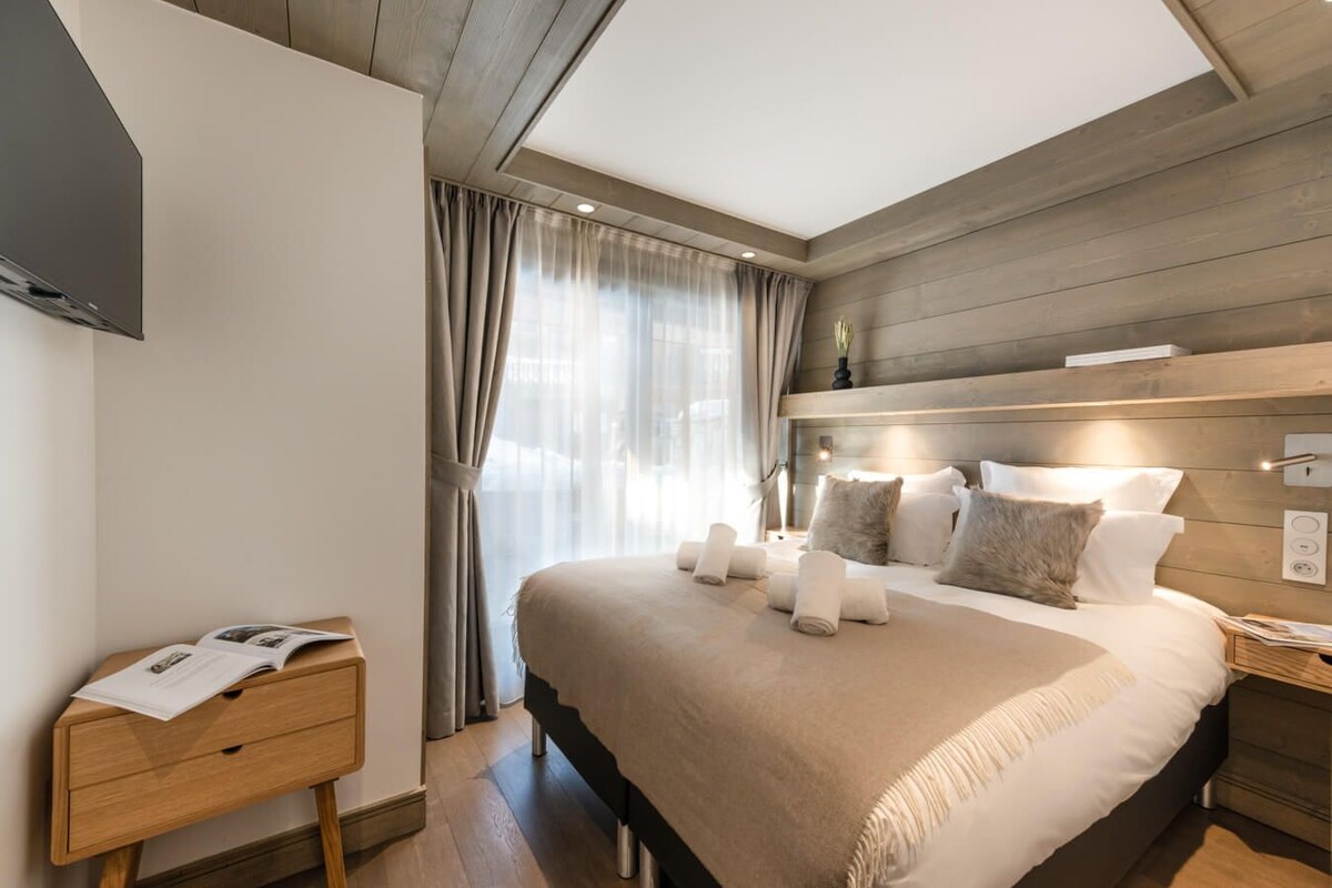 A modern hotel room with a large bed, a bedside table with a magazine, a wooden headboard, and a window with curtains.