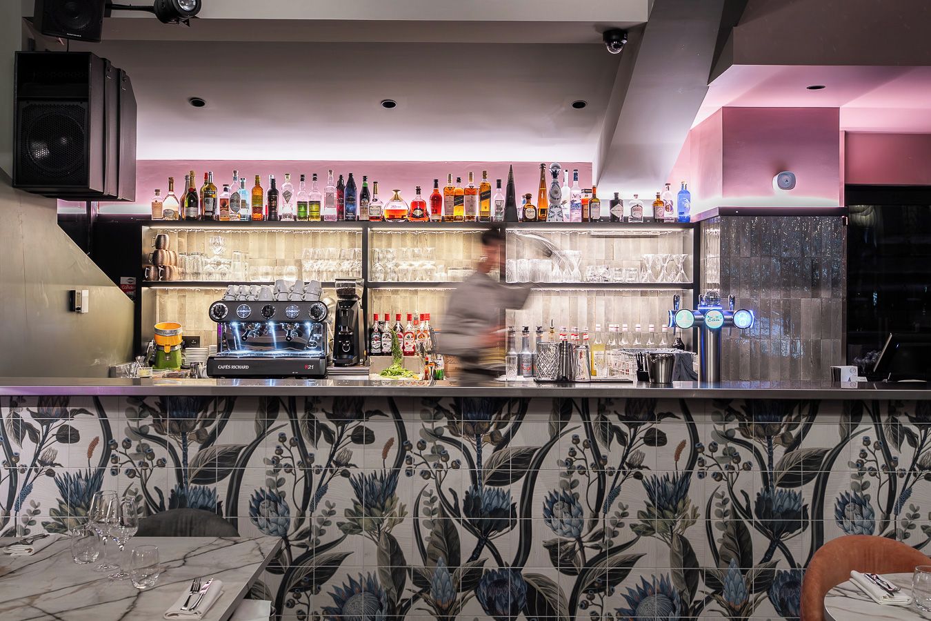 A bar area with a coffee machine, shelves of liquor bottles, and a tiled floral backsplash.