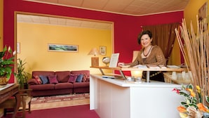 A reception desk with a woman seated behind it, a sofa, a lamp, and a plant in a room with red walls. - Villa Ferrari Hotel Garni (San Maurizio d'Opaglio)
