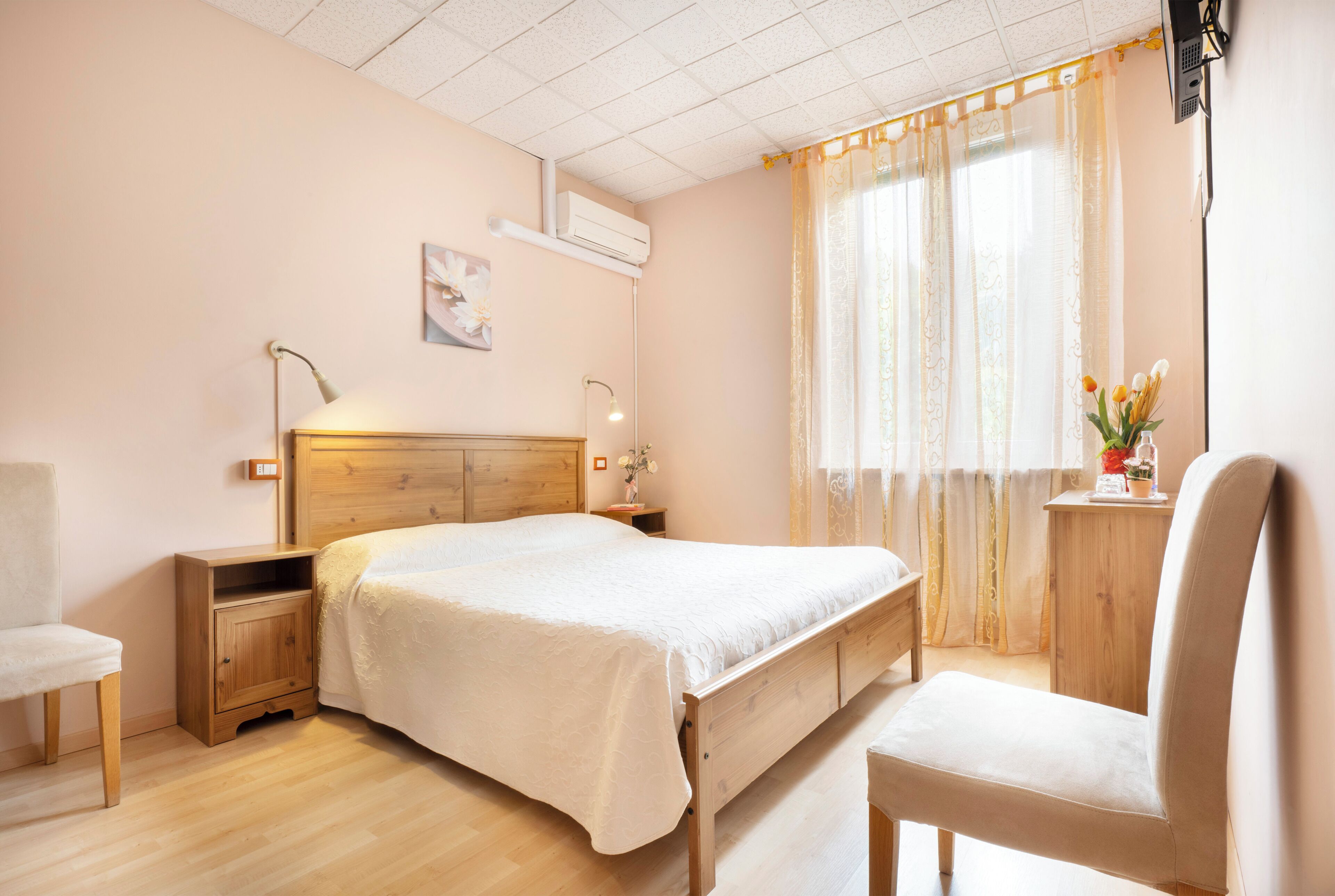 A bedroom with a wooden bed, bedside tables, a chair, a small desk with a vase of flowers, and a window with curtains.