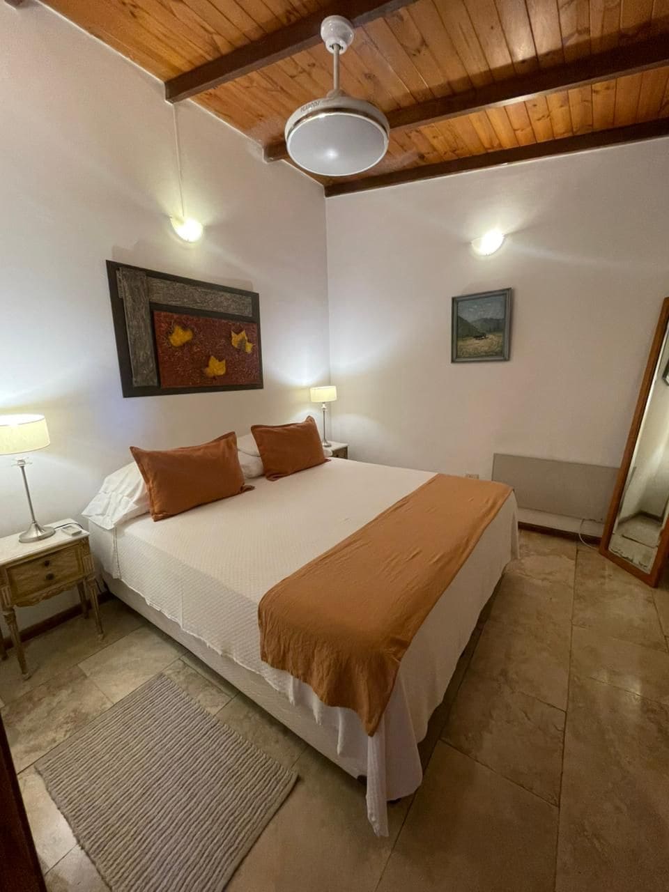 Un dormitorio con cama, mesitas de noche, lámpara, ventilador de techo y obras de arte en las paredes.