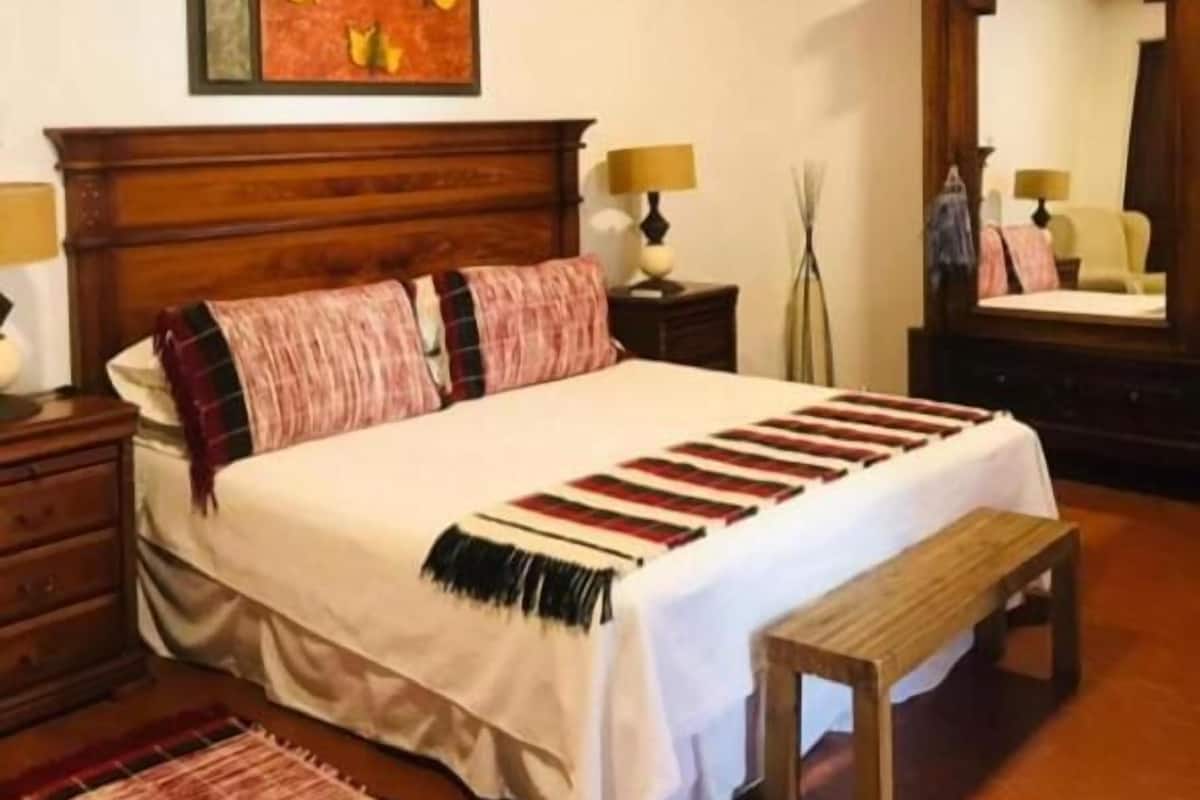 A bedroom with a wooden bed, a bench, a nightstand, a lamp, a mirror, and a framed picture on the wall.