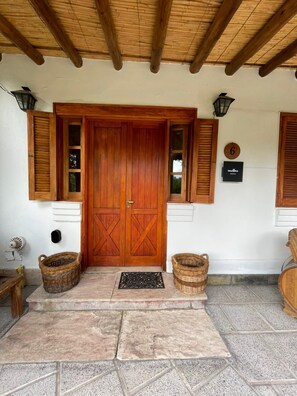 Una puerta de madera con un diseño de cruz, flanqueada por dos ventanas con contraventanas, y un área de entrada con pavimento de piedra.