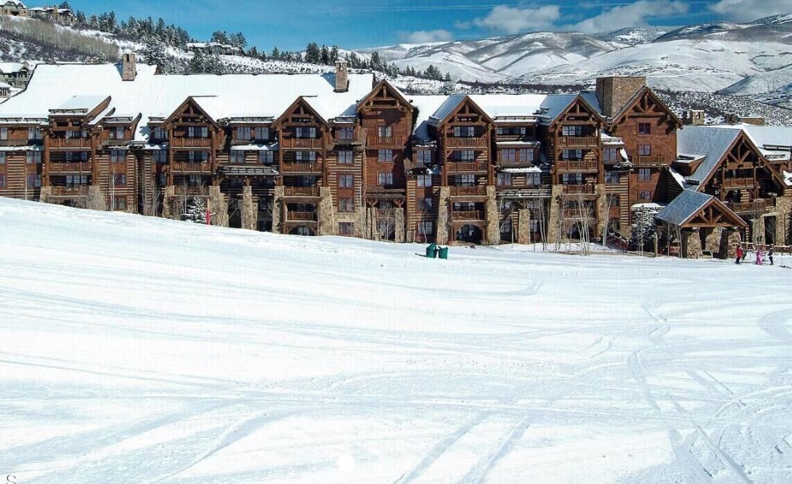 A large lodge with multiple wooden buildings, a ski slope in the foreground, and snow-covered mountains in the background.