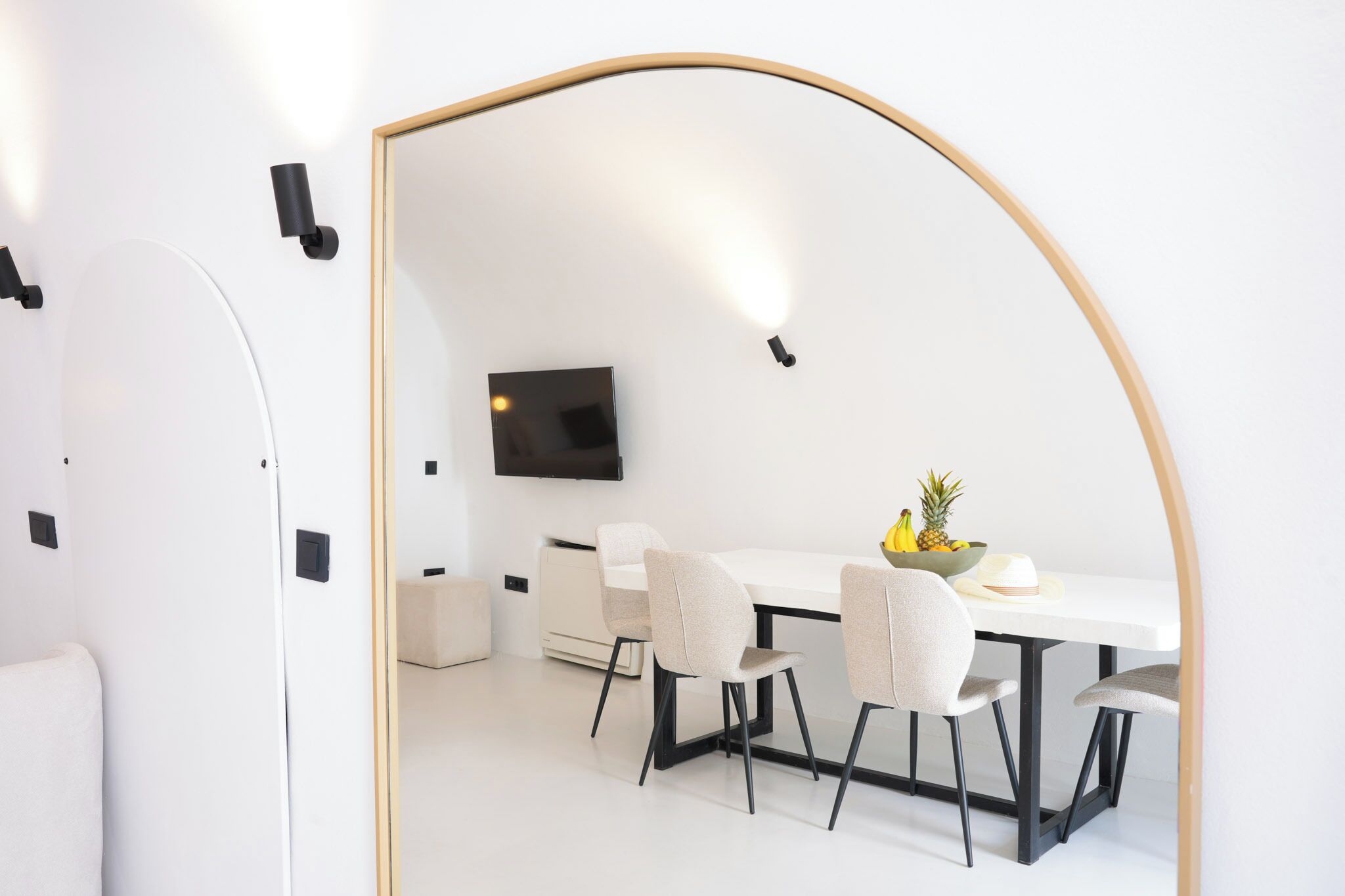 A modern dining area with a white table, four chairs, a flat-screen TV mounted on the wall, and a fruit bowl on the table.