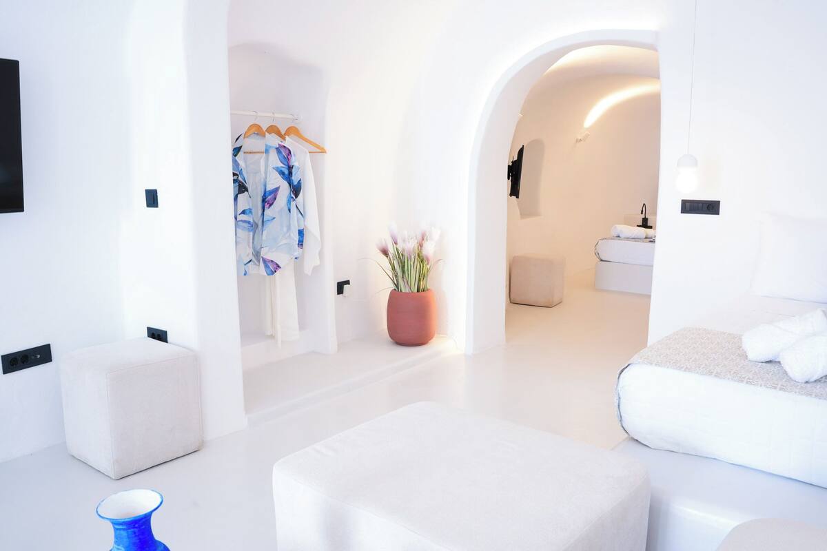 A modern hotel room with a minimalist design, featuring a large bed, a seating area, a bathroom, and a hallway leading to another room.