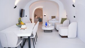 A modern living room with a white sofa, a dining table with chairs, and a coffee table. There is a TV mounted on the wall and a doorway leading to another room. - Eros suites Oia (Oia)