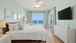 Un dormitorio con cama, ventilador de techo, televisor, ventana con vista al océano y cómoda.