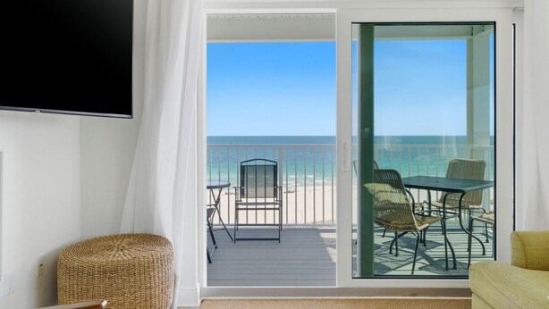 Un balcón con vista a la playa, un televisor en la pared y un reposapiés de mimbre.