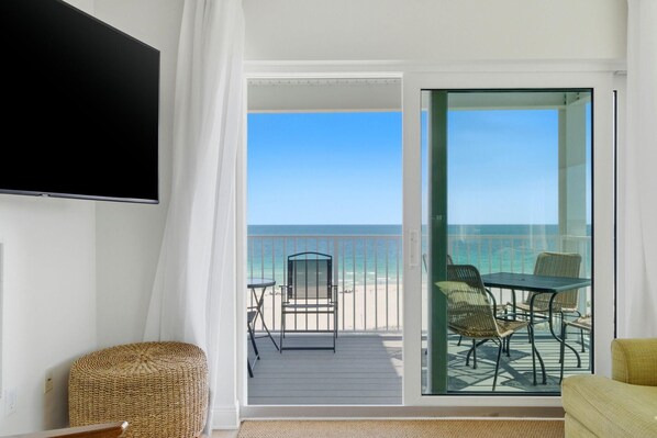 Ein Balkon mit Blick auf den Strand, ein an der Wand befestigter Fernseher und ein Korbsessel.