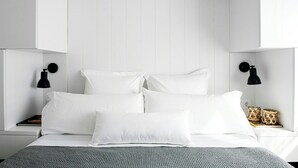 Una cama bien hecha con ropa de cama y almohadas blancas, dos lámparas de noche negras y una cesta tejida en la mesita de noche.