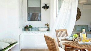 Una habitación iluminada por el sol, con una mesa de madera, un banco, una ventana con contraventanas, un lavabo y una lámpara fijada a la pared.