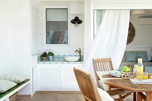 A sunlit room with a wooden table, a bench, a window with shutters, a sink, and a wall-mounted light fixture. - Designed Costa Brava flat with panoramic sea views (Girona)