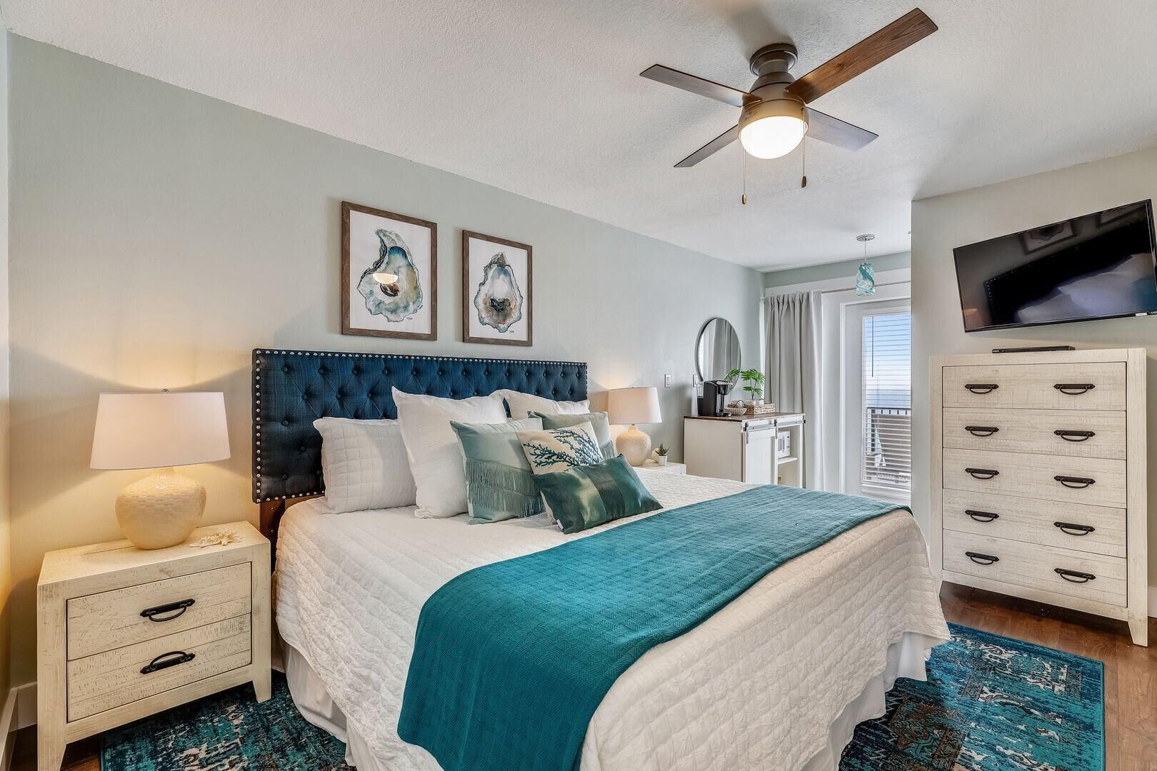 A bedroom with a bed, nightstands, a dresser, a ceiling fan, artwork, a television, and a window with curtains.