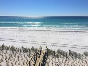 Ein Strand mit einer Holzpromenade, die zum Meer führt, Sanddünen und einem strahlend blauen Himmel.