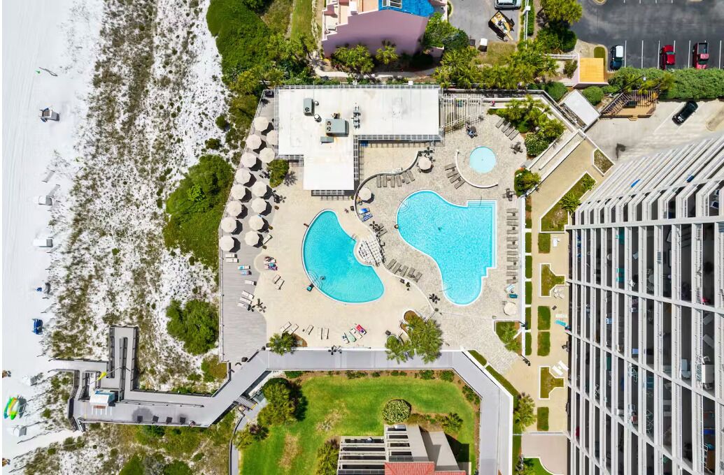 Luftaufnahme eines Hotels mit Poolbereich, Parkplatz und Strand.