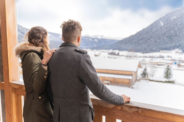A couple standing by a wooden balcony overlooking a snowy landscape with mountains in the background.