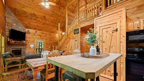 Ein Holzblockhaus-Interieur mit Kamin aus Stein, einem Esstisch, Stühlen, einer Küchenzeile mit Herd und einem an der Wand montierten Fernseher.