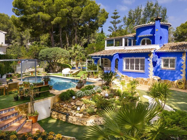 Une maison bleue avec une piscine, entourée de verdure et d’un mur de pierres.