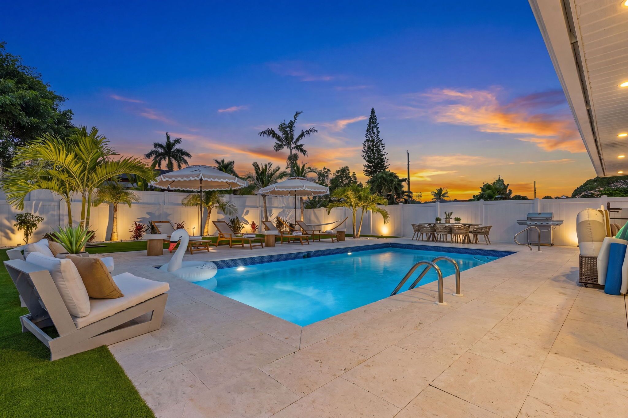 A swimming pool with lounge chairs and umbrellas, surrounded by greenery and a sunset view.