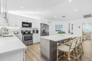 A modern kitchen with white cabinetry, stainless steel appliances, a central island with bar stools, and a large window. - A Touch of Heaven • Heated Pool • Near Beach (Naples)
