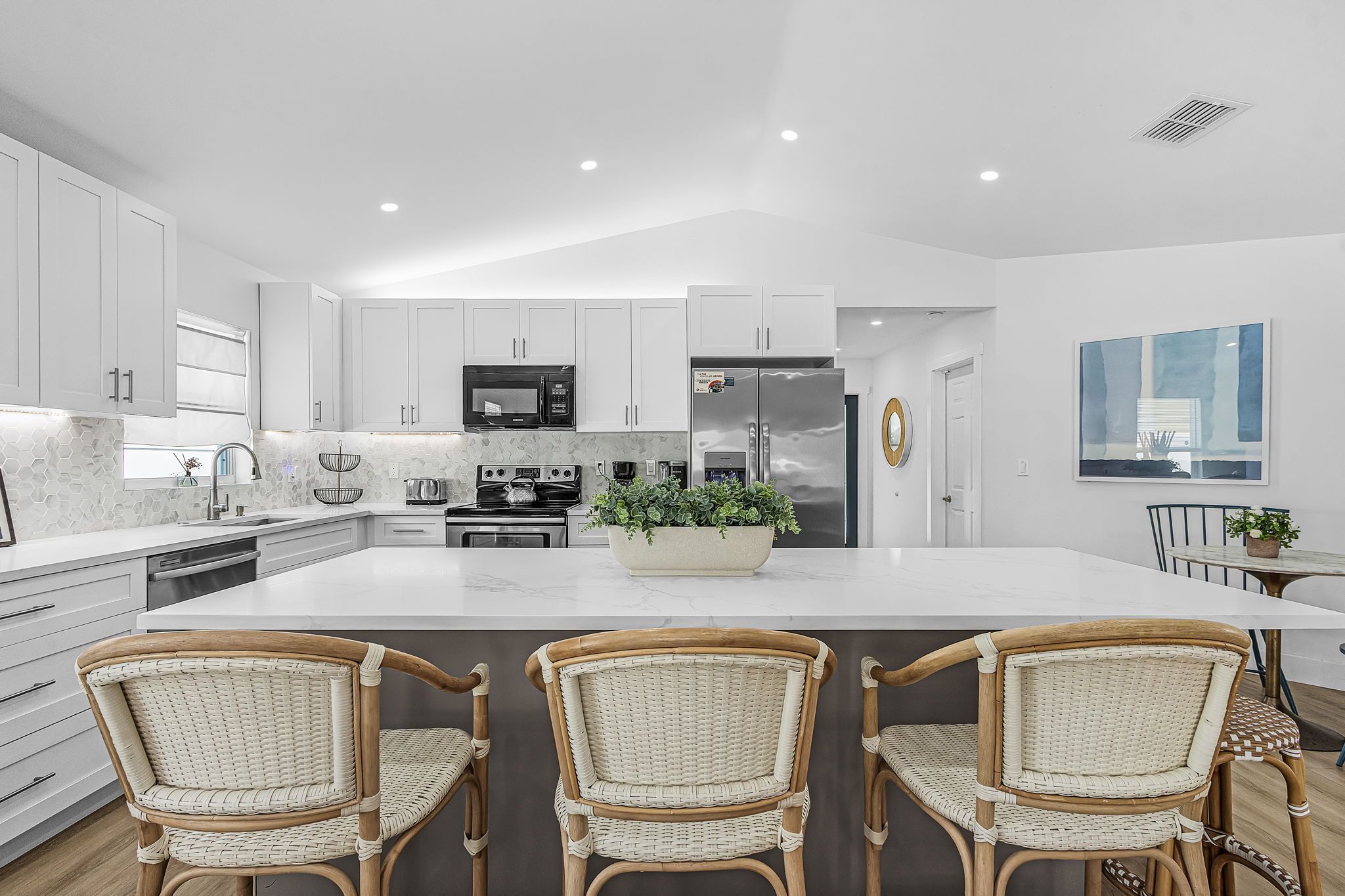 A modern kitchen with white cabinets, a central island, and three wicker chairs.