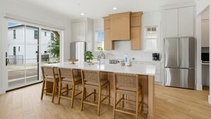 A modern kitchen with a central island, bar stools, stainless steel appliances, and a sliding glass door leading to an outdoor area. - The Tannin House - Meeting House Square 7 (Orange Beach)