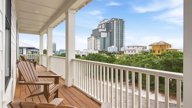 A wooden deck with chairs, a white railing, and a view of a cityscape with modern buildings. - The Tannin House - Meeting House Square 7 (Orange Beach)