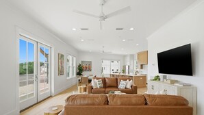 A spacious living area with a large brown leather sofa, a flat-screen TV mounted on the wall, and a dining area in the background. - The Tannin House - Meeting House Square 7 (Orange Beach)