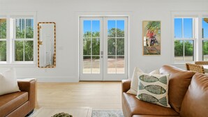 A spacious living room with a brown leather sofa, a coffee table, and a large window with a view of greenery. - The Tannin House - Meeting House Square 7 (Orange Beach)