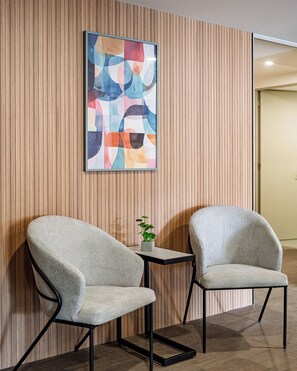 Ein Zimmer mit zwei grauen Sesseln, einem kleinen Tisch mit Pflanze und einem farbenfrohen abstrakten Gemälde an einer Holzwand.