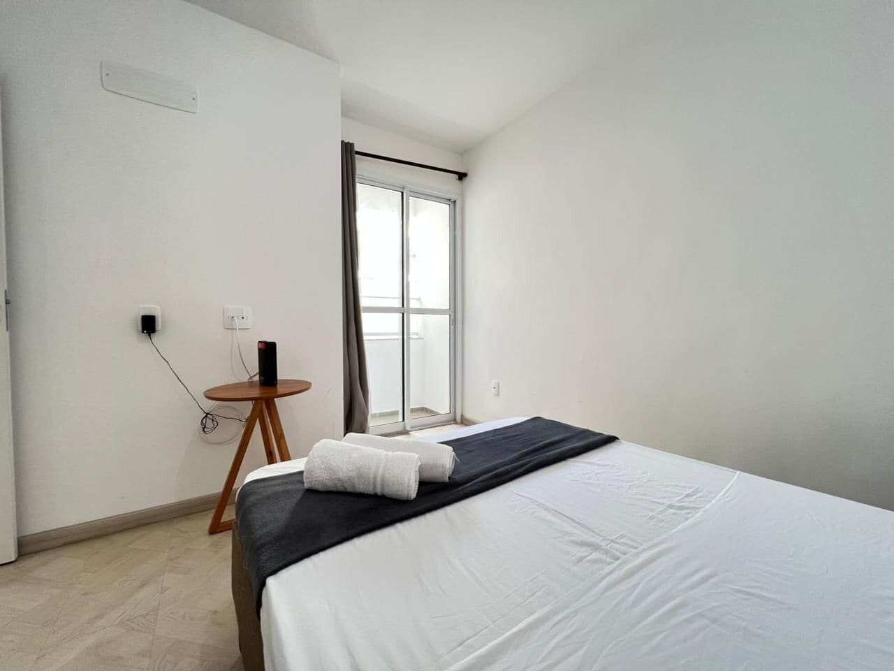 A hotel room with a bed, a small table with a chair, a window with curtains, and a wall socket.