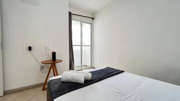 A hotel room with a bed, a small table with a chair, a window with curtains, and a wall socket.